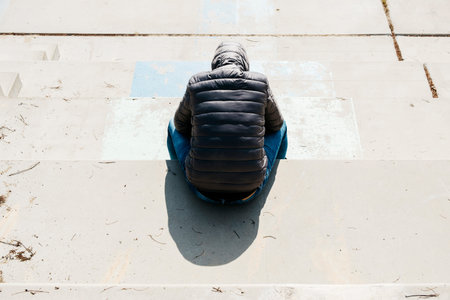 a young caucasian man, seen from behind, wearing jeans and a gray hooded jacket, curled up sitting...