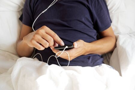 closeup of a young caucasian man in pajamas lying down in bed using his smartphone, with the earbuds plugged to itの写真素材