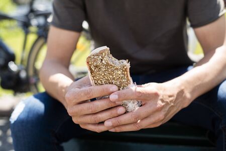 closeup of a young caucasian man, wearing denim jeans and a t-shirt, eating a turkey sandwich while sitting on a park, next to some bicycles in the backgroundの写真素材