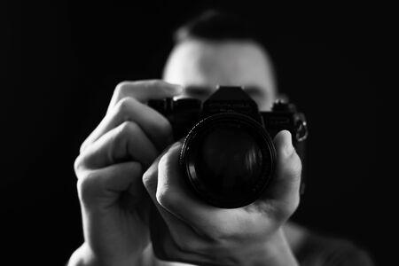 closeup of a young man looking through the viewfinder of a reflex camera, pointing to the observer, about to press the shutter button, in black and whiteの写真素材