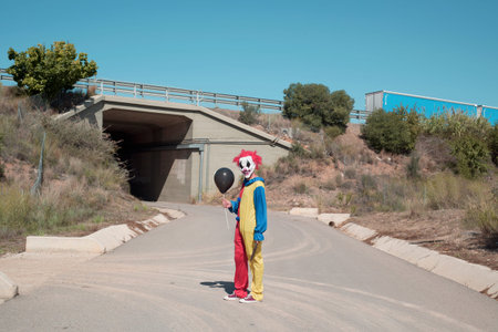 a scary clown wearing a colorful yellow, red and blue costume outdoors, holding a black balloon in...