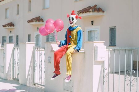 a creepy clown wearing a colorful yellow, red and blue costume, holding a bunch of red balloons in his hand, sitting in front of a row of housesの写真素材