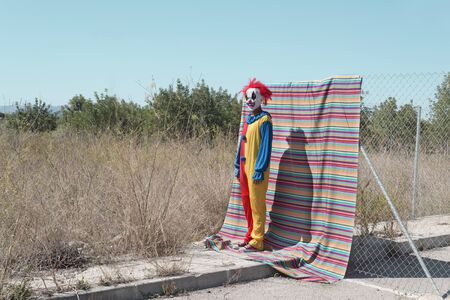 a scary clown, in a colorful yellow, red and blue costume, standing in front of a fence, with a colorful striped piece of cloth hanging from it, and an abandoned natural landscape in the backgroundの写真素材
