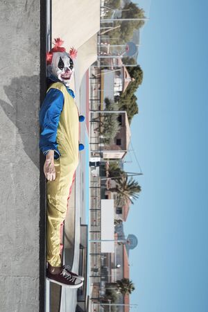 a scary clown, wearing a colorful yellow, red and blue costume, lying down in an outdoor public sports complexの写真素材