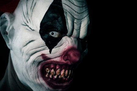 closeup of a scary evil clown with  white eyes, bloody teeth and a threatening look staring at the...