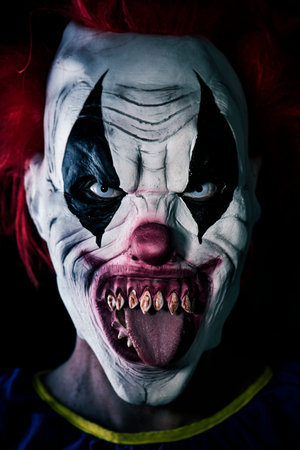 closeup of a scary evil clown with red hair, white eyes, bloody teeth, sticking out his tongue...