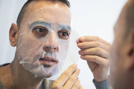 Closeup of a handsome caucasian man applying a bio-cellulose sheet mask to his own faceの写真素材