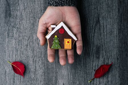 high angle view of a young caucasian man with a christmas cookie in the shape of a gingerbread house in his hand, against a gray wooden surfaceの写真素材