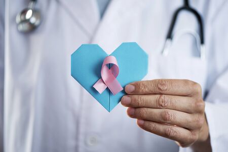 closeup of a young caucasian doctor man, wearing a white coat, holding a blue origami heart and a pink ribbon in his handの写真素材