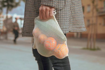 closeup of a man in the street carrying a textile reusable mesh bag, used to buy groceries in bulk, full of fruit and vegetables, as a measure to reduce plastic pollutionの写真素材
