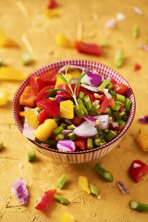 closeup of a bowl with a mix of different raw chopped vegetables, such as asparagus, onion, or yellow and red bell pepper, on a golden textured surfaceの写真素材
