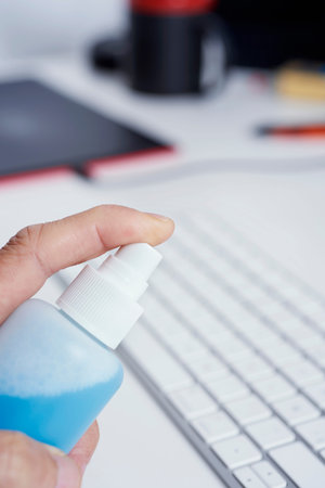 Closeup of a caucasian man disinfecting a computer keyboard by spraying a blue sanitizer from a...