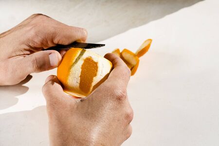 Closeup of a caucasian man peeling an orange with a table knife, on a white tableの写真素材