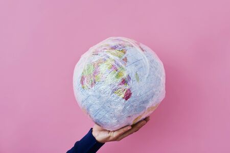 a man holds a world globe wrapped in plastic, depicting the plastic contamination or the protection against the epidemic infectious diseases or the air pollution, on a pink backgroundの写真素材