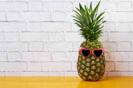 a pineapple wearing a pair of pink heart-shaped sunglasses on yellow surface against a white brick...