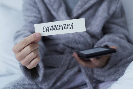 closeup of a man, wearing a gray robe staying at home or a hotel room, showing a note with the word cuarentena, quarantine in spanish written in itの写真素材