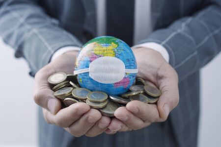 businessman holding a world globe with a protective mask on top of a handful of euro coins, depicting the economic consequences of the coronavirus pandemic or of the pollution and climate changeの写真素材
