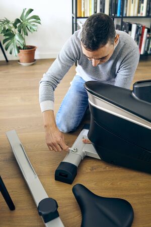 A caucasian man assembling the different pieces of a stationary bicycle in the living room of his houseの写真素材