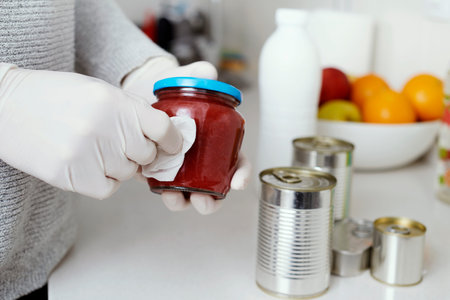 A man in the kitchen, wearing latex gloves, cleaning food cans and jars, freshly purchased, with a...