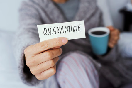 A man, in pajamas staying at home or a hotel room, showing a note with the word quarantine written in itの写真素材