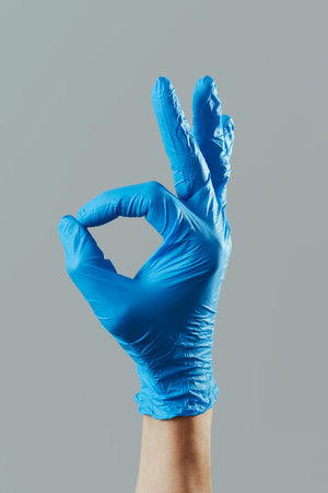 closeup of the hand of a man, wearing blue surgical gloves, doing the OK sign against a gray...
