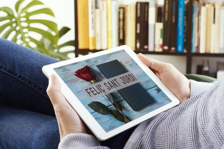 man watching, in a tablet, a picture of a rose, a book and the text Happy Saint Georges Day written in Catalan, made by myself, when it is tradition to give red roses and books in Catalonia, Spainの写真素材