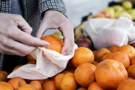 closeup of a man shopping at a greengrocer putting some mandarin oranges in a textile reusable mesh bag, as a measure to reduce plastic pollutionの写真素材