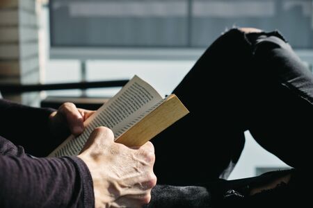 closeup of a young caucasian man, in casual wear, reading a book sitting in an outdoors armchair on the balconyの写真素材