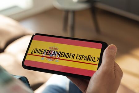 closeup of a young caucasian man, wearing casual clothes, sitting in a chair indoors, having his smartphone in his hand with the text do you want to learn Spanish written in Spanish in its screenの写真素材