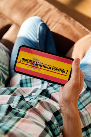 closeup of a young caucasian man, wearing casual clothes, sitting in a chair indoors, having his smartphone in his hand with the text do you want to learn Spanish written in Spanish in its screen