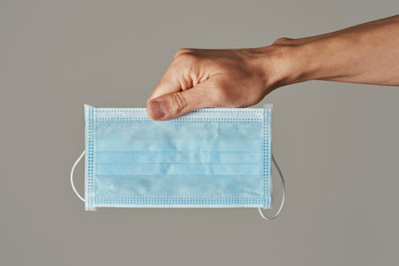 closeup of a young caucasian man with a surgical mask in his hand, on a gray background