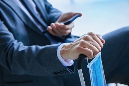 a businessman, in a gray suit, sitting in the waiting hall of an airport or a train station, using his smartphone and holding a surgical mask while is leaning his hand on the handle of his suitcaseの写真素材