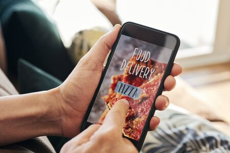 closeup of a young caucasian man, in the living room, having his smartphone in his hand with the text food delivery pizza in its screenの写真素材