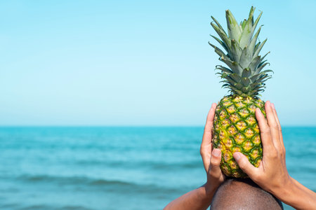 closeup of a man seen from behind on the beach, in front of the sea, holding a pineapple on his head, with some blank space on the leftの写真素材