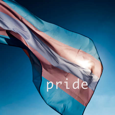 closeup of a transgender pride flag waving on the blue sky, moved by the wind, with the sun in the background, and the word prideの写真素材