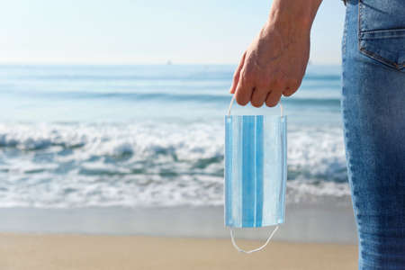 closeup of a young caucasian man in jeans, seen from behind, standing on the beach holding a blue surgical mask in his hand, as is taking a breather of wearing it, with the sea in the backgroundの写真素材