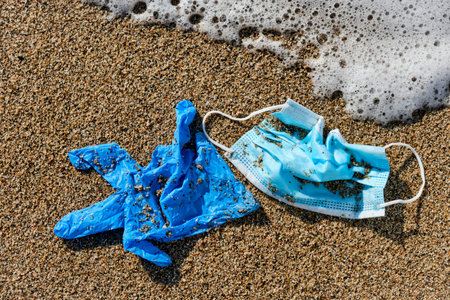 Closeup of a blue used surgical mask and a blue latex glove thrown on the wet sand of the seashore...
