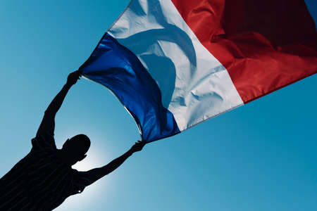 closeup of the silhouette of a young man holding a French flag waving on the wind against the blue sky, in backlitの写真素材