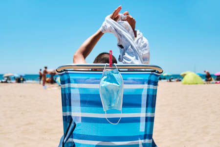 closeup of a caucasian man, sitting in a deck chair on the beach, seen from behind, taking off  his T-shirt while his surgical mask is hanging from the chair, pinned with a clothespinの写真素材