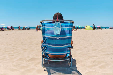 closeup of a caucasian man, sitting in a deck chair on the beach, seen from behind, and his surgical mask hanging from the chair, as he is taking a breather of wearing it or sunbathingの写真素材