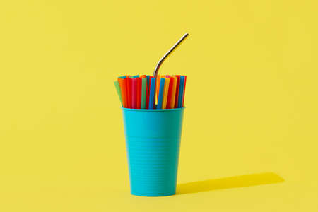 a reusable stainless steel drinking straw standing out from some disposable plastic drinking straws of different colors in a blue vase, on a yellow background with some blank space around themの写真素材