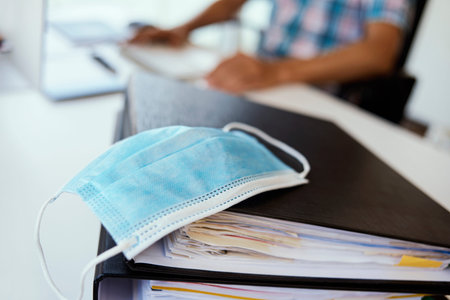 closeup of a blue disposable face mask on an office desk and a young man using a computer in the...