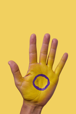 closeup of the intersex flag painted in the palm of the hand of a young person, against a yellow...