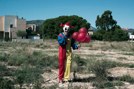 a creepy clown wearing a yellow, red and blue costume, holding a bunch of red balloons in his hand, standing in a vacant lotの写真素材
