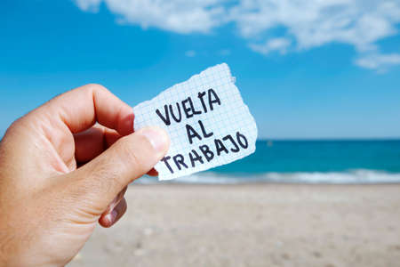 closeup of the hand of a young caucasian man on the beach, in front of the ocean, holding a piece of paper with the text back to work written in spanishの写真素材