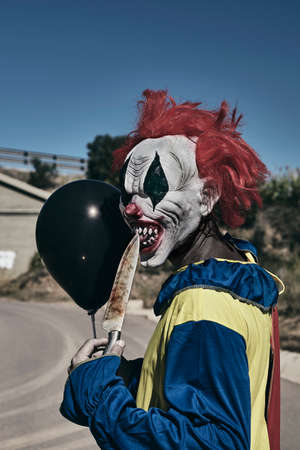 closeup of a scary evil clown outdoors, holding a black balloon in one hand and bringing the tip of a big knife to his mouth, while staring at the observer with a threatening lookの写真素材