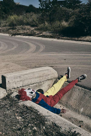 a scary clown, wearing a yellow, red and blue costume, lying down face up on the gutter of a secondary roadの写真素材