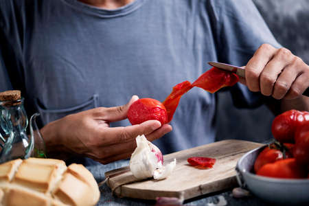 closeup of a young caucasian man, wearing a gray t-shirt, peeling some scalded tomatoes sitting at a kitchen table, next to a loaf of bread and a cruet with olive oilの写真素材