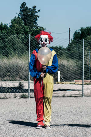 a scary clown wearing a yellow, red and blue costume, holding a golden balloon in his hands, standing on the asphalt outdoorsの写真素材