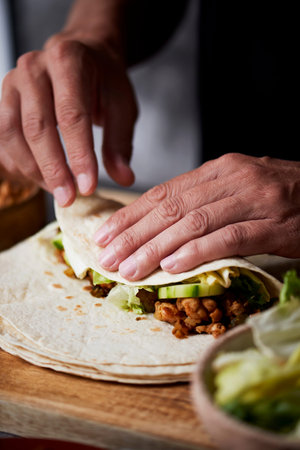 closeup of a young caucasian man preparing a durum or a burrito, with chicken meat, lettuce and...